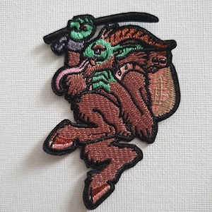 May include: Embroidered patch depicting a brown, green, and pink creature with a black border. The creature has a long tongue and is holding a black object. The patch is set against a white backdrop.