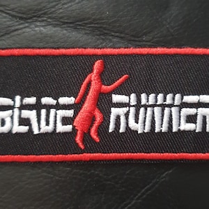 Puede incluir: Un parche bordado negro y rojo con una silueta blanca de una persona corriendo sobre una valla blanca con el texto "BLADE RUNNER" en blanco.