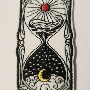 May include: An embroidered patch featuring an hourglass with a sun and moon inside. The top of the hourglass shows a red sun with rays and clouds. The bottom of the hourglass shows a yellow crescent moon with clouds and a black starry sky.