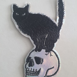 May include: An embroidered patch showing a black cat on a skull. The cat has detailed fur and intense eyes. The skull has a pearlescent finish. This patch is suitable for jackets, bags, or other fabric items, and can be sewn or ironed on.