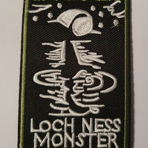 May include: A black and green embroidered patch with the text "Cryptozoology Tracking Society" and "Loch Ness Monster" in white. The patch also features a white embroidered image of the Loch Ness Monster.
