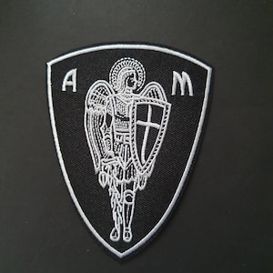 Op de afbeelding: Zwarte geborduurde patch met een witte omtrek in de vorm van een schild. Het schild heeft een witte afbeelding van een engel met vleugels, een zwaard en een schild met een kruis. De letters "A" en "M" staan aan weerszijden van de engel.