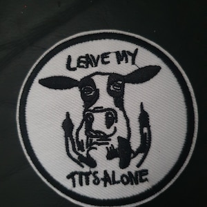 Op de afbeelding: Ronde geborduurde patch met een zwart-witte koe die haar middelvinger opsteekt. De tekst "LEAVE MY TITS ALONE" is in zwart geborduurd op een witte achtergrond met een zwarte rand.