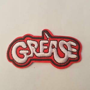 以下が含まれることがあります： 「Grease」という単語がスタイリッシュなフォントで刺繍された赤と白の刺繍パッチ。パッチは車の形をしています。