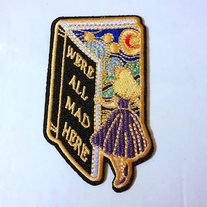 Puede incluir: Un parche bordado para planchar que presenta un libro con el texto "We're All Mad Here" y una imagen de dibujos animados de Alicia de Alicia en el País de las Maravillas saliendo del libro. El fondo es un cielo nocturno estrellado con una luna creciente.