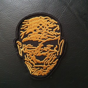 Peut inclure: Écusson brodé avec un motif de visage jaune doré sur fond noir. Le patch a une bordure noire. Le design est détaillé et complexe, adapté à une application sur des vêtements ou des accessoires.