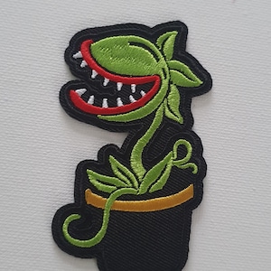 May include: An embroidered patch depicting a carnivorous plant with a toothy mouth, in a black pot with a gold rim. The plant is green and red.
