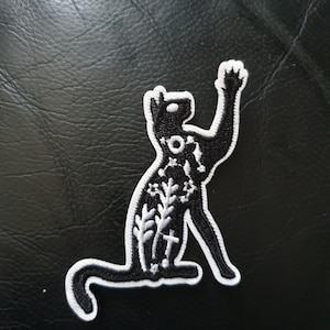 May include: Black and white embroidered iron-on patch of a cat with floral details. The cat is sitting up with its paw raised.