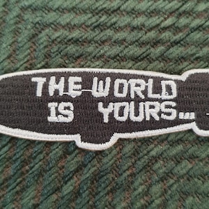 Peut inclure: Patch brodé noir et blanc en forme de dirigeable avec le texte "THE WORLD IS YOURS..."