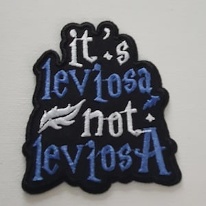 Könnte beinhalten: Schwarzer gestickter Aufnäher mit weißer und blauer Schrift, die "it's leviosa not leviosa" lautet.