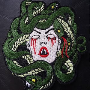 May include: Green embroidered patch of Medusa with snakes for hair, white face, red lips, and red tears.