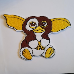 May include: Embroidered patch of a cartoon gremlin. The patch has a brown body, white accents, yellow ears and feet, and large brown eyes. The gremlin is holding a small object.