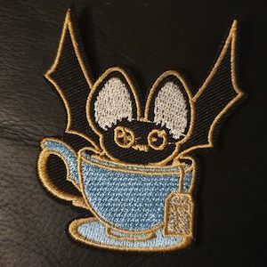 May include: An embroidered black bat with white ears sitting in a blue teacup with a gold rim and a tea bag hanging from the side.