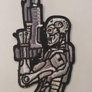 Może przedstawiać: Naszywka haftowana w kolorze srebrnym i czarnym przedstawiająca robota trzymającego dużą broń. Robot ma czerwone oko i szczegółowy wzór.