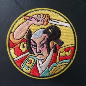 Könnte beinhalten: Gestickter Aufnäher mit einem Samurai-Krieger, der ein Schwert über seinem Kopf hält. Der Krieger trägt einen roten und grünen Kimono vor einem gelben Hintergrund. Der Aufnäher ist rund und hat einen gelben Rand.