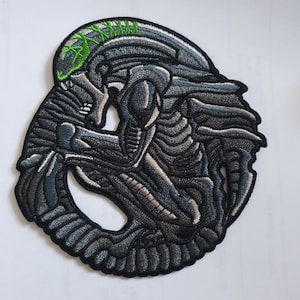 May include: A black and grey embroidered patch of an alien creature with a green spine. The alien is curled up in a foetal position.