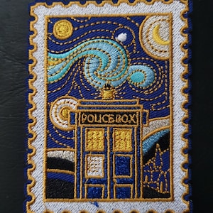 Peut inclure: Écusson brodé représentant une cabine de police bleue avec le texte "POLICE BOX" sur un fond de ciel étoilé tourbillonnant, inspiré de "La Nuit étoilée" de Van Gogh. L'écusson a une bordure blanche avec une garniture dorée.