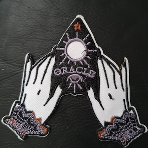 Pode incluir: Patch bordado com um design celestial. Apresenta mãos brancas, um triângulo preto com a palavra "ORACLE" e um sol. O patch tem uma estrela e um detalhe de olho, adequado para jaquetas, bolsas ou outros artigos de tecido.