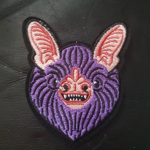 May include: Purple and pink embroidered iron-on patch of a bat with a toothy grin.