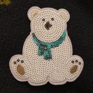 May include: A white polar bear with a teal blue sequined scarf. The bear is made of sequins and is sitting down.
