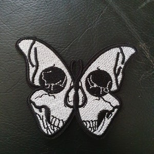 Puede incluir: Un parche bordado en blanco y negro de una mariposa con alas con forma de calavera.