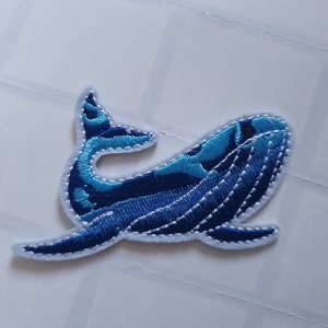 Puede incluir: Parche de ballena bordado en azul con costuras blancas. La ballena está mirando hacia la derecha y tiene un diseño estampado en azul y blanco.