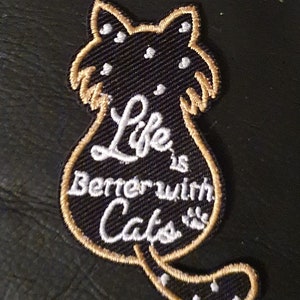 Könnte beinhalten: Ein gestickter Aufnäher in Form einer Katze mit dem Text "Life is Better with Cats" in Weiß auf schwarzem Hintergrund.