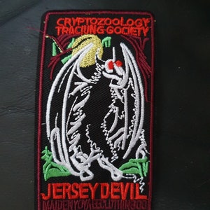 May include: A black and white embroidered patch with a red border. The patch features a stylised image of the Jersey Devil, a mythical creature with wings and red eyes. The text "CRYPTOZOOLOGY TRACING SOCIETY" is above the image, and the text "JERSEY DEVIL MAIDENYKRAECLOTHINGCO" is below the image.