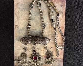 May include: A silver necklace with a long, cylindrical pendant with intricate designs. The pendant has a red stone and hangs from three silver tassels. The necklace also has a silver chain and a silver whistle.