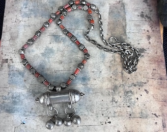 May include: A silver necklace with a pendant featuring a small, cylindrical box with bells. The necklace is made of silver beads and coral-colored beads.