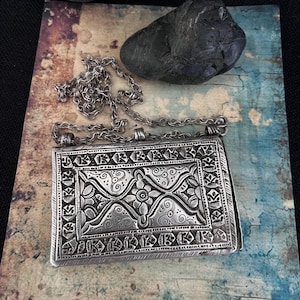 May include: A silver pendant with an intricate floral design, featuring a chain and a rectangular shape. The pendant has a detailed, embossed pattern.
