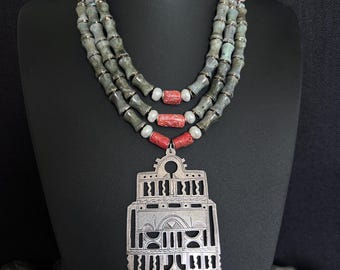 May include: A silver pendant necklace with a geometric design hangs from a triple strand of gray and green beads with red coral accents.
