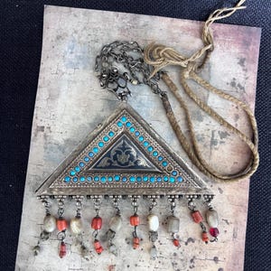 May include: A triangular silver pendant with turquoise stones and a black enamel design. The pendant is attached to a brown cord and a silver chain.