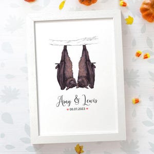 May include: A white framed print with a black and white illustration of two bats hanging upside down and kissing. The text "Amy & Lewis 08.07.2023" is printed below the bats.