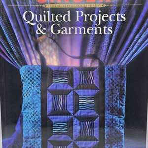 May include: A softcover book titled SINGER Sewing Reference Library Quilted Projects & Garments. The cover features a blue and purple quilted tote bag with a geometric pattern, set against a backdrop of draped blue and purple fabric.