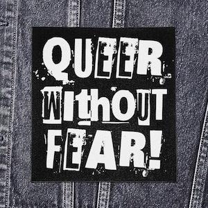 Puede incluir: Parche cuadrado negro con el texto blanco "QUEER Without FEAR!" en una fuente desgastada. El parche está colocado sobre una chaqueta vaquera oscura.