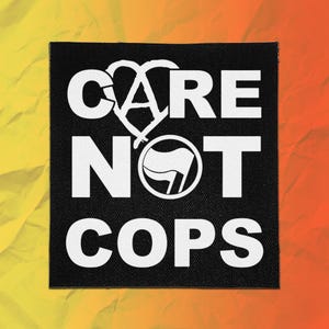May include: A square black fabric patch featuring the white text CARE NOT COPS. The letter A in CARE is stylized as a heart shape, and the letter O in NOT is replaced by a circular icon containing a small flag symbol in white on a black background.