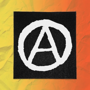 May include: A square fabric patch featuring a black background with a white hand-drawn style letter A enclosed within a circle. The design is centered on the patch, which has a woven texture and clean, straight edges throughout the entire square shape.
