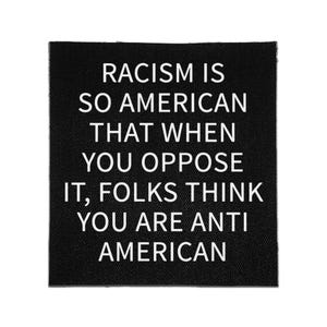 May include: A square black fabric patch featuring white text that reads: RACISM IS SO AMERICAN THAT WHEN YOU OPPOSE IT, FOLKS THINK YOU ARE ANTI AMERICAN. The patch has a visible woven texture and clean, straight edges on all four sides.