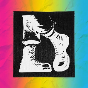 Op de afbeelding: Een vierkante stoffen patch met een contrastrijke zwart-witte grafische illustratie. Het ontwerp toont een paar benen in een broek met omgeslagen pijpen en veterschoenen, weergegeven in een strakke sjabloonstijl met opvallende zwarte vlakken.