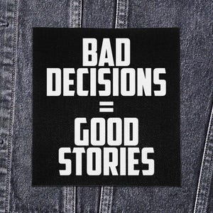 May include: Black square patch with the text "BAD DECISIONS = GOOD STORIES" in bold white letters. The patch is set against a dark denim jacket.