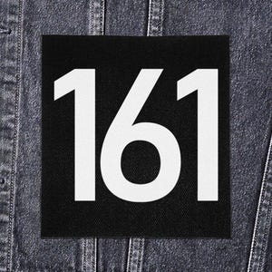 May include: A black square patch with the white number "161" prominently displayed. The patch is set against a denim jacket background, creating a contrast between the bold number and the textured fabric.