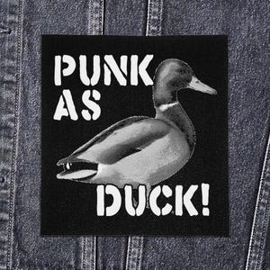 May include: Black and white patch featuring a duck illustration. The text "PUNK AS DUCK!" is printed in white, bold, capital letters. The patch is square and appears to be sewn onto a denim jacket.