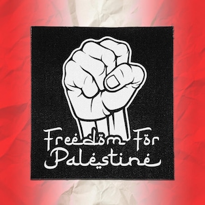 Puede incluir: Un parche de tela cuadrado de color negro con un gráfico blanco de un puño levantado. Debajo del puño, el texto "Freedom För Palestine" está impreso en una fuente blanca estilizada. El parche tiene una textura tejida visible en el fondo.