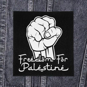 Puede incluir: Parche cuadrado negro con un puño cerrado blanco. El parche incluye las palabras "Freedom For Palestine" en una fuente estilizada. El parche está sobre un fondo de chaqueta vaquera.