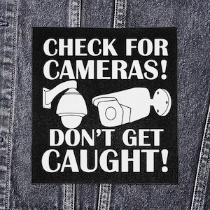 Op de afbeelding: Zwart vierkantige patch met witte tekst en illustraties van beveiligingscamera's. De tekst luidt "CHECK FOR CAMERAS! DON'T GET CAUGHT!". Twee beveiligingscamera-ontwerpen zijn afgebeeld. De patch wordt getoond op een spijkerjasje.