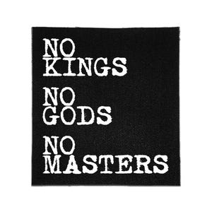Op de afbeelding: Een vierkante zwarte stoffen patch met de in wit gedrukte tekst NO KINGS NO GODS NO MASTERS verdeeld over drie regels. De letters hebben een verweerd, getextureerd uiterlijk tegen de effen zwarte achtergrond van het geweven textiel.