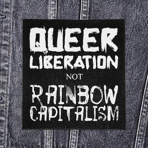 Puede incluir: Parche cuadrado negro con texto blanco: "QUEER LIBERATION NOT RAINBOW CAPITALISM". El texto es en negrita y desgastado. El parche está sobre una chaqueta vaquera.