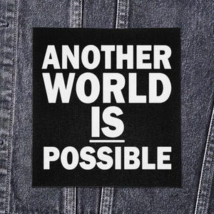 May include: A black square patch with the white text "ANOTHER WORLD IS POSSIBLE." The patch is displayed on a dark denim jacket. The text is in a bold, sans-serif font.
