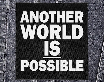 May include: A black square patch with the white text "ANOTHER WORLD IS POSSIBLE." The patch is displayed on a dark denim jacket. The text is in a bold, sans-serif font.